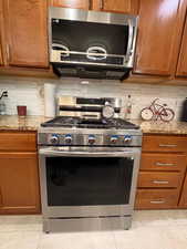 Kitchen featuring stainless steel appliances, brown cabinets, light tile patterned floors, light stone countertops, and tasteful backsplash