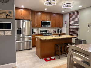 Kitchen featuring light stone countertops, tasteful backsplash, appliances with stainless steel finishes, a kitchen breakfast bar, and brown cabinetry