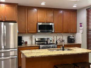 Kitchen featuring light stone counters, appliances with stainless steel finishes, brown cabinetry, backsplash, and recessed lighting