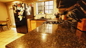 Kitchen featuring dark countertops, backsplash, black refrigerator with ice dispenser, light tile patterned flooring, and extractor fan
