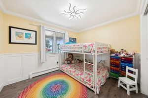 Bedroom featuring a baseboard heating unit, crown molding, a wainscoted wall, dark wood finished floors, and a decorative wall