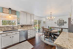 Kitchen with hanging light fixtures, stainless steel dishwasher, dark wood-style flooring, light stone counters, and backsplash