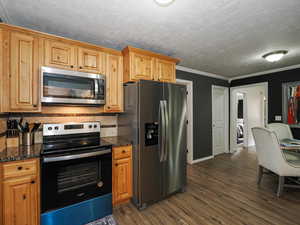Kitchen with appliances with stainless steel finishes, dark wood-style floors, dark stone counters, decorative backsplash, and ornamental molding