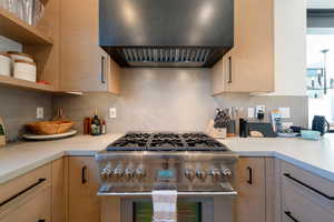 Kitchen with wall chimney range hood, high end stainless steel range oven, open shelves, backsplash, and light brown cabinets