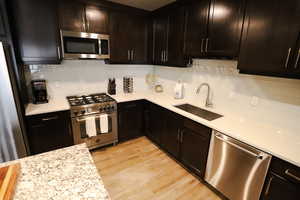 Kitchen with appliances with stainless steel finishes, backsplash, light stone countertops, light wood finished floors, and dark brown cabinets