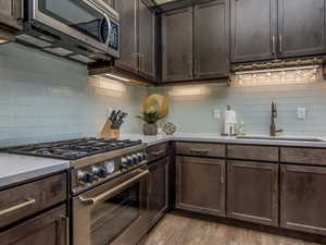 Kitchen with dark brown cabinets, stainless steel appliances, tasteful backsplash, light wood-style floors, and light stone counters