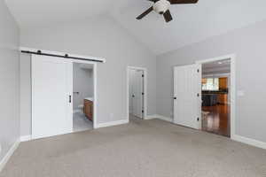 Unfurnished bedroom featuring a barn door, light carpet, ensuite bathroom, high vaulted ceiling, and a ceiling fan