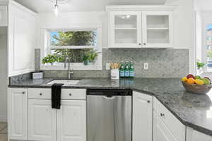 Kitchen featuring white cabinets, dishwasher, dark stone counters, glass insert cabinets, and tasteful backsplash
