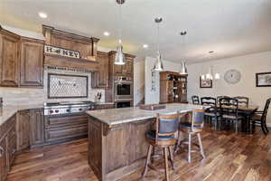 Kitchen with light stone countertops, hanging light fixtures, dark wood-type flooring, stainless steel appliances, and tasteful backsplash