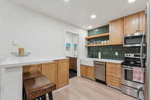 Kitchen with open shelves, appliances with stainless steel finishes, brown cabinets, light wood-style flooring, and backsplash