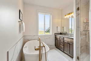Full bath featuring double vanity, a freestanding bath, tile walls, wainscoting, and a marble finish shower