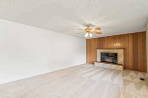 Unfurnished living room featuring wood walls, light colored carpet, a textured ceiling, ceiling fan, and a brick fireplace