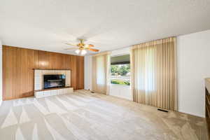 Unfurnished living room featuring a fireplace, light carpet, wooden walls, a textured ceiling, and a ceiling fan