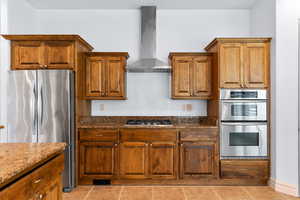 Kitchen with appliances with stainless steel finishes, wall chimney exhaust hood, brown cabinetry, and dark stone countertops