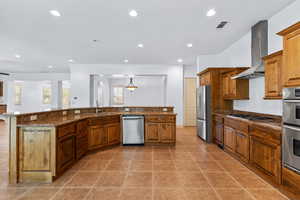 Kitchen with brown cabinetry, appliances with stainless steel finishes, wall chimney range hood, dark stone counters, and recessed lighting