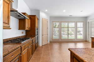 Kitchen featuring brown cabinets, dark stone counters, light tile patterned floors, wall chimney range hood, and recessed lighting