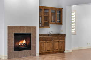 Bar featuring a tiled fireplace, dark stone counters, brown cabinetry, glass insert cabinets, and light tile patterned floors