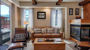Carpeted living area with a tiled fireplace, beam ceiling, and plenty of natural light