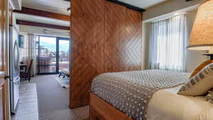 Carpeted bedroom featuring wood walls, access to exterior, freestanding refrigerator, tile patterned flooring, and beamed ceiling