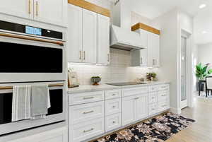 Kitchen featuring double oven, decorative backsplash, light countertops, premium range hood, and light wood-style floors