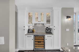 Indoor dry bar featuring wine cooler, decorative backsplash, light stone countertops, white cabinetry, and glass insert cabinets