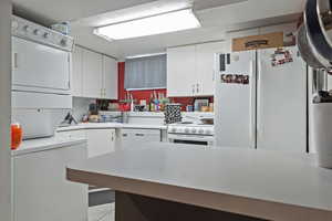 Kitchen with white cabinetry, light countertops, white appliances, and estacked washer and dryer