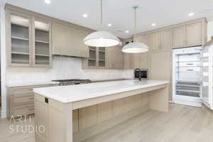 Kitchen with a large island, tasteful backsplash, light brown cabinets, glass insert cabinets, and hanging light fixtures