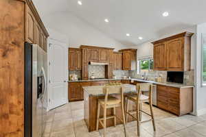 Kitchen with brown cabinets, lofted ceiling, light stone counters, light tile patterned flooring, and stainless steel refrigerator with ice dispenser