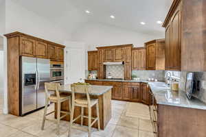 Kitchen with brown cabinets, light stone counters, stainless steel fridge, decorative backsplash, and high vaulted ceiling