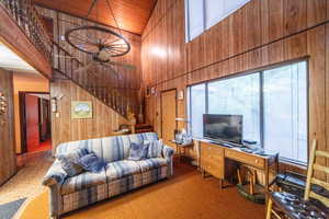 Carpeted living room featuring wood walls, high vaulted ceiling, a ceiling fan, stairs, and wood ceiling