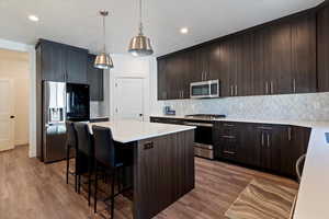 Kitchen with decorative backsplash, dark brown cabinets, appliances with stainless steel finishes, a kitchen island, and hanging light fixtures