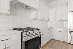 Kitchen featuring stainless steel gas range, wall chimney range hood, white cabinetry, tasteful backsplash, and freestanding refrigerator
