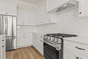 Kitchen with appliances with stainless steel finishes, white cabinetry, backsplash, wall chimney exhaust hood, and light wood-style flooring