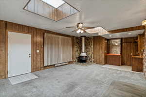 Unfurnished living room featuring wooden walls, carpet, a wood stove, a ceiling fan, and a textured ceiling