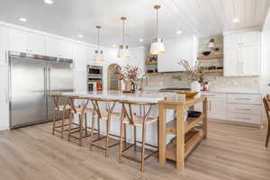 Kitchen with open shelves, built in appliances, white cabinetry, hanging light fixtures, and light stone counters