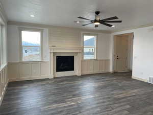 Unfurnished living room featuring crown molding, dark wood-type flooring, a fireplace, ceiling fan, and a decorative wall