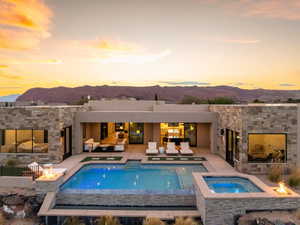 Back of property at dusk with an outdoor fire pit, a mountain view, an in-ground hot tub, a patio, and an outdoor stone fireplace