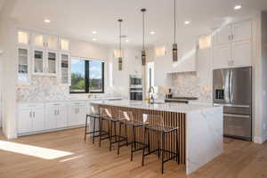 Kitchen featuring glass insert cabinets, appliances with stainless steel finishes, an island with sink, white cabinets, and decorative light fixtures