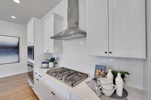 Kitchen featuring wall chimney exhaust hood, white cabinets, appliances with stainless steel finishes, decorative backsplash, and light wood-type flooring