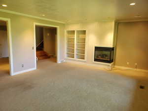 Unfurnished living room featuring recessed lighting, crown molding, a glass covered fireplace, carpet, and built in shelves