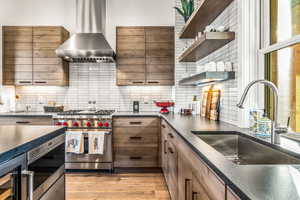 Kitchen with open shelves, wall chimney range hood, appliances with stainless steel finishes, backsplash, and light wood finished floors