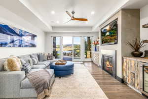 Living area featuring ceiling fan, wood finished floors, a glass covered fireplace, a raised ceiling, and recessed lighting
