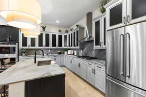 Kitchen featuring appliances with stainless steel finishes, light stone countertops, decorative light fixtures, a kitchen island with sink, and wall chimney exhaust hood