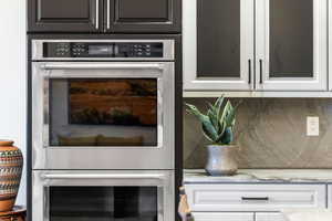 Kitchen view of double oven, decorative backsplash, light stone countertops, and white cabinets