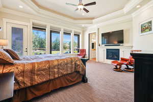 Bedroom with light colored carpet, a raised ceiling, ensuite bath, a glass covered fireplace, and ornamental molding