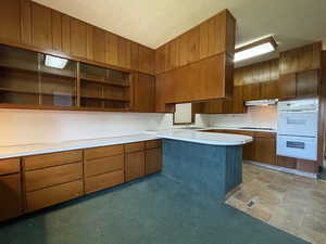 Kitchen with light countertops, brown cabinetry, white double oven, a peninsula, and under cabinet range hood