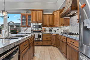 Kitchen featuring backsplash, appliances with stainless steel finishes, custom exhaust hood, pendant lighting, and brown cabinetry