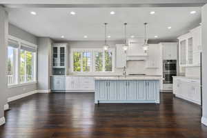 Main level kitchen with glass fronted cabinets, hanging light fixtures, huge kitchen island with sink, stainless steel double oven, and backsplash