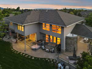 Back of property at dusk featuring stucco siding, a patio, a shingled roof, a lawn, and stone siding - and one of the most private backyards you'll ever see!