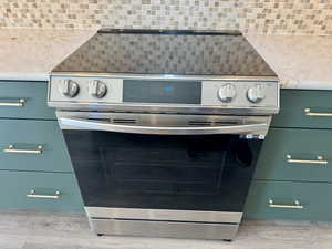 Detailed view of stainless steel electric range, green cabinetry, light stone counters, light wood finished floors, and backsplash